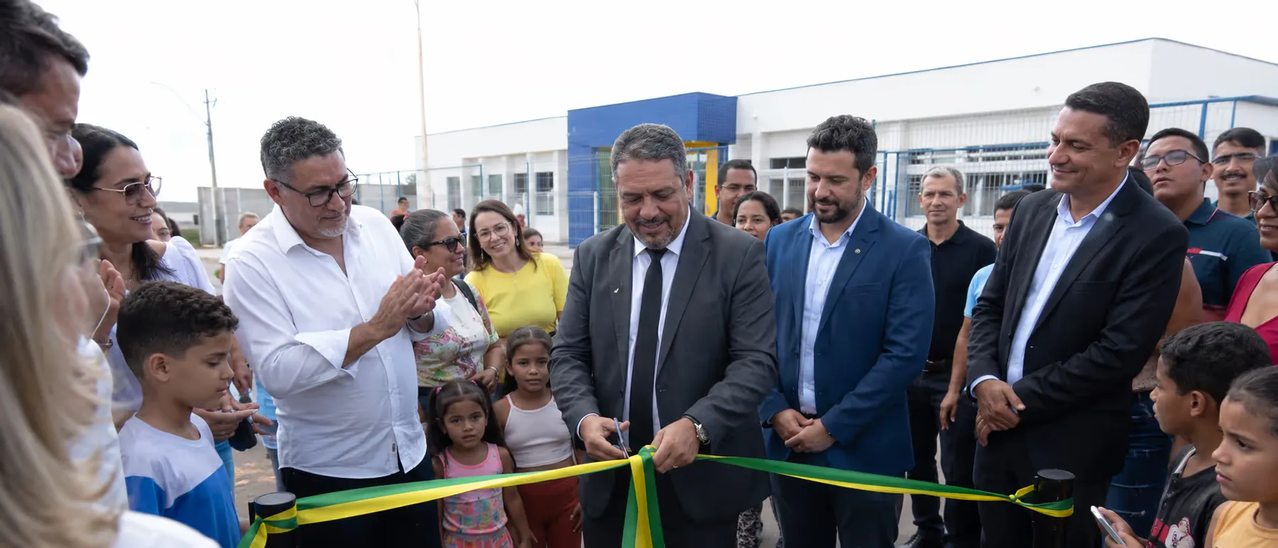 Cerimônia de inauguração ao ar livre: autoridades cortam uma fita verde e amarela cercadas por moradores, adultos e crianças