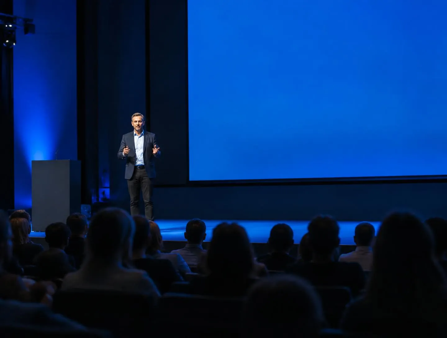 Professor falando em um auditório universitário diante de uma tela azul, com a plateia em primeiro plano